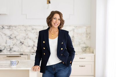 Woman with short wavy brown hair standing in a bright kitchen, smiling and wearing a navy blazer, white top, and jeans, with a patterned mug on the counter beside her.