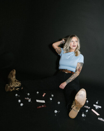 woman in black pants and light blue shirt sits against a black wall with small disco balls scattered around her. 