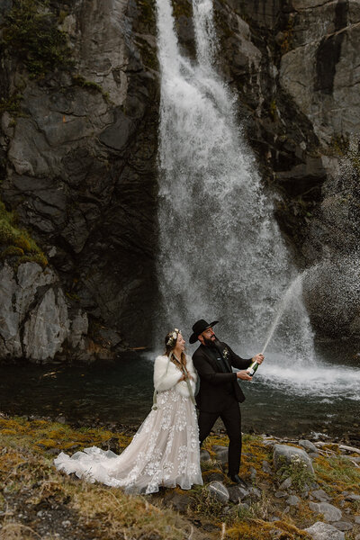 Couple eloping pops champagne with Alaska waterfall in background.
