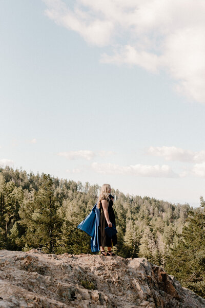 Senior holding her gown and staring off while standing on a rock on the top of a mountain