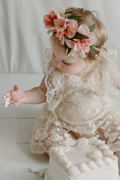 Baby girl looking at her hand with white icing on it and cake sitting in front of her.