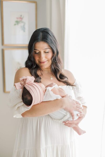 A mother wearing a white dress stands by a window holding her newborn baby who is wearing a pink bow.