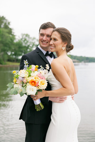 man in tux hugging girl in white dress smiling standing in front of the water