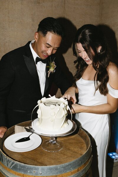 Bride and Groom are smiling while cutting their wedding cake on top of a wine barrel at Seven Branches Venue and Inn in Sonoma California