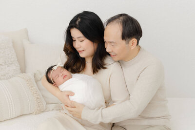 New mom and dad dressed in neutral colors are holding their newborn baby in their arms and looking down at her. 