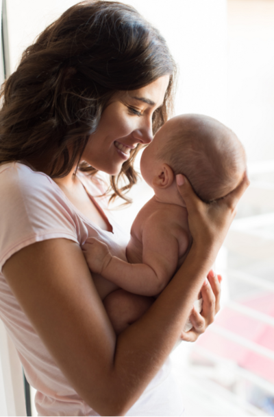 A loving moment at a Texas postnatal retreat: a new mom, dressed in a pastel pink blouse, tenderly holds her beautiful baby girl and leans in to kiss her cheeks.