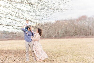 boy sits on dad's shoulders and parents smile at each other during family photo session with Sara Sniderman Photography in Wellesley Massachusetts