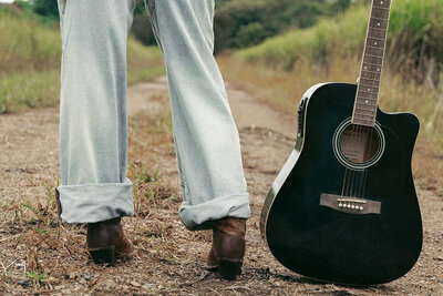 Person wearing jeans and brown shoes stands on a dirt path next to a black acoustic guitar, with grass and trees in the background—a scene that captures the creative spirit of Sweet Pea Ink Creative’s brand design.
