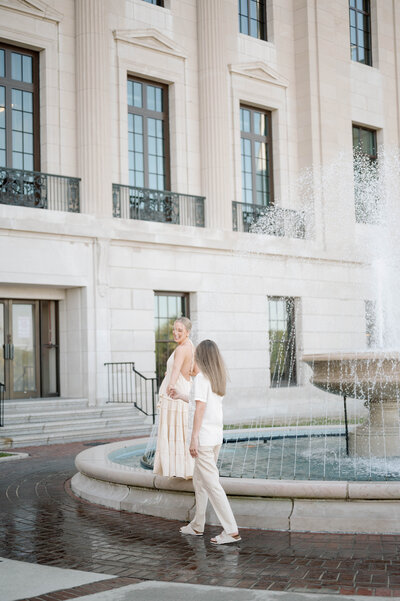 Wearing all white, a woman leans against a concrete wall in downtown Raleigh with her fiancé to her side. He is also leaning against the wall, looking at her.