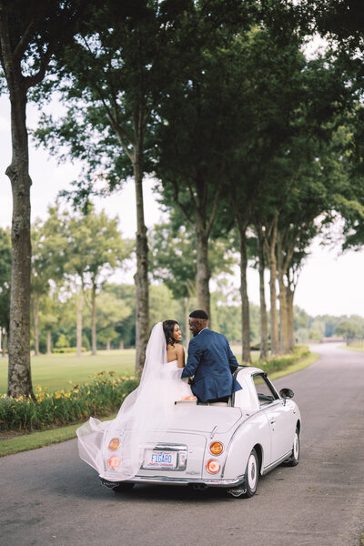 Bride and groom sitting on top of vintage car among a row of trees