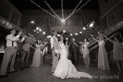A bride and groom stand smiling under string lights at night, surrounded by their wedding party holding sparklers in celebration. Everyone is dressed formally and appears joyful.