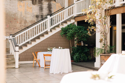 An outdoor courtyard set for an event, with round tables covered in white cloths, wooden chairs, floral centerpieces, potted plants, and string lights overhead.