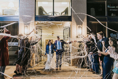 A bride and groom walk out of a building at night, smiling as guests on both sides throw white streamers in celebration. The scene is festive, with people cheering and taking photos.