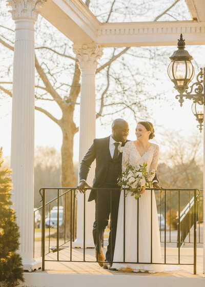 A newlywed couple standing at a small railing smiling at each other at sunset 