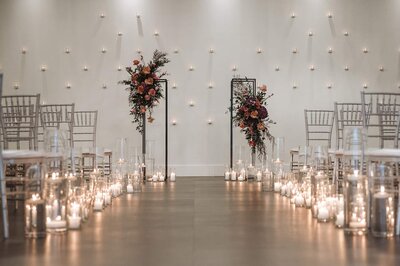 A candlelit ceremony aisle leads to two modern floral pillars at SoHo63, a Chandler wedding and event venue. Surrounded by silver chairs and a backdrop of floating wall candles, the bold floral arrangements add a vibrant pop of color to the modern, romantic setting.
