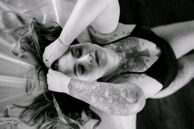 black and white boudoir photography with woman who had many tattoos on the edge of a bed with her hands in her hair with her eyes closed