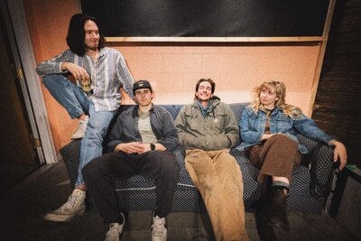 Four people sitting casually on a couch in a relaxed, indoor setting, dressed in casual, vintage-inspired clothing with a laid-back, playful vibe.  Representing Good Morning June vendor profile for the Crush Collective wedding show.
