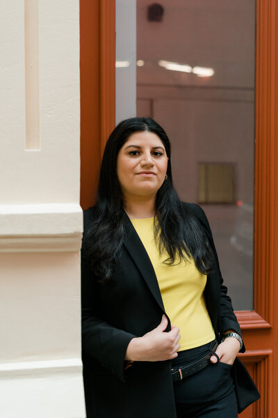 woman standing in doorway leaning against wall while holding one side of her jacket and the other hand is in pocket