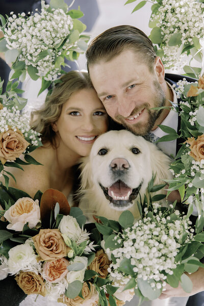 A couple poses with their dog, a wreath of orange and white flowers surrounding them.