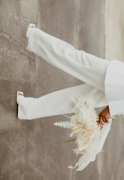 person in white suite and heels walking sideways holding white bouquet