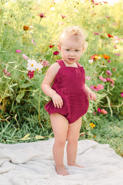 A toddler in a red romper looks straight at the camera with a serious look. She is standing on a white blanket in a wildflower field.