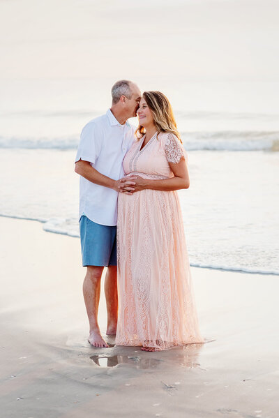 Expectant mother in pink dress with husband standing by the water in Atlantic Beach, FL.