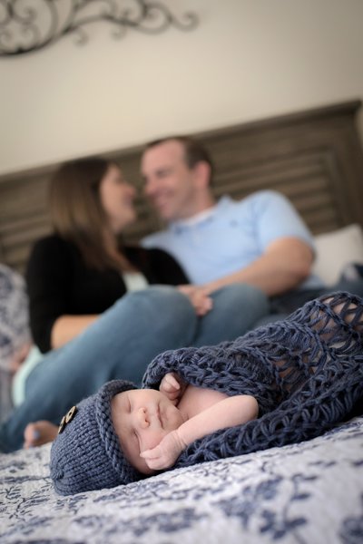 A sleeping newborn wrapped in a blue knitted blanket and hat is in the foreground. In the background, a couple sits on a bed, smiling warmly.