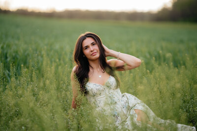 woman sitting on the ground in a field while touching her hair and wearing a dress