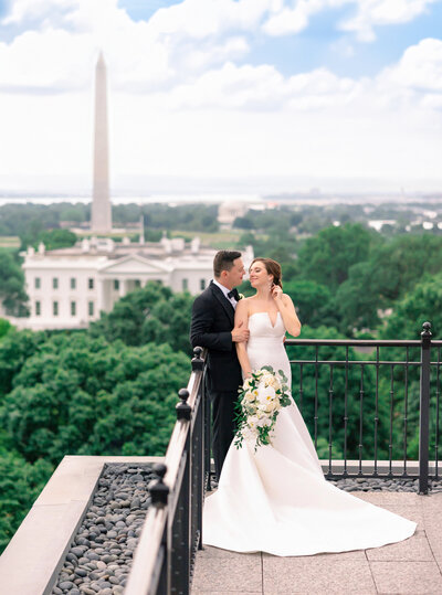 A bride and groom stand on a rooftop overlooking the White House and Washington Monument. The bride is in an elegant white dress holding a bouquet of flowers, while the groom wears a tuxedo.
