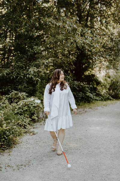 In the background of the greener of the Stanley Park trail and stone pathway, Anne, an Asian woman, is walking with her white cane. She is looking over her left shoulder and smiling. She is wearing a white knee length dress and suede espadrilles.