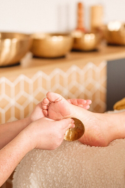 Gros plan sur des mains tenant un bol kansu en bronze posé sur la plante des pieds lors d'un massage ayurvédique à Challans, Vendée, Padabhyanga.