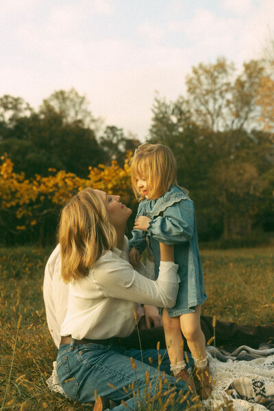 A mother sitting on a blanket reaching to pick up her young daughter who is wearing a cute demin dress. 