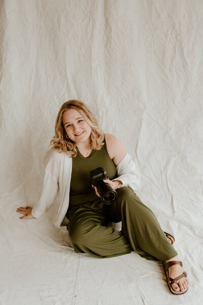 A person with curly hair smiles while holding a camera, sitting on a beige sofa. They wear a green checkered sweater and ripped jeans, creating a cozy, creative atmosphere.