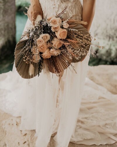 A close-up of a bridal bouquet featuring peach roses, dried palm leaves, and various dried foliage. The bouquet is being held by a bride wearing a lace wedding gown. Keywords: luxury destination weddings, bridal bouquet, destination wedding planning.