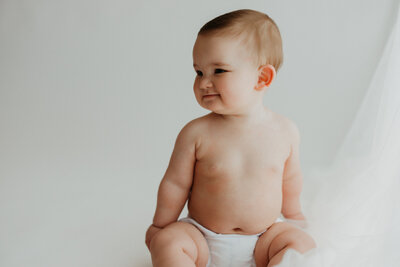 six month old baby wearing a diaper cover sits nice and tall on a white rug during his milestone Mini session in Denver.
