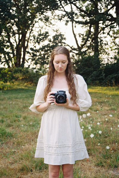 Lifestyle photographer, Heather, of Among the Oaks holds a film camera and looks down at it while standing in a field.