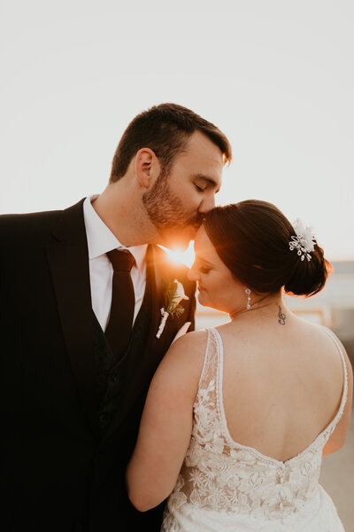 Groom kissing bride's forehead with sunset flare peeking through their faces