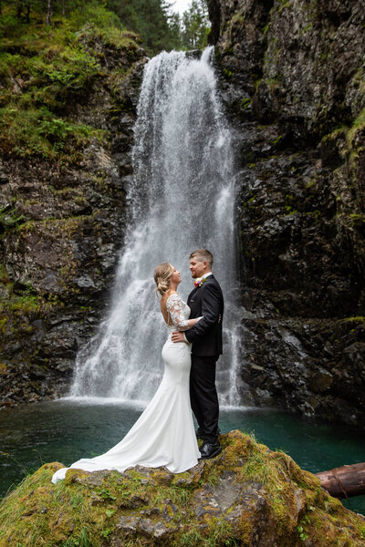 Groom playfully lifts bride in a mountain valley, both laughing and surrounded by lush alpine scenery.