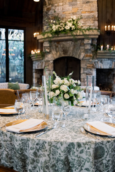 stone fireplace with greenery and flowers behind circular table with white flower centerpiece and glasses on the table
