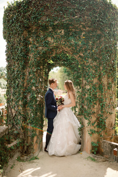 bride and groom standing facing each other in an ivy covered garden tower archway. A rainbow colored sunflare cuts across in the lower left side of the photograph