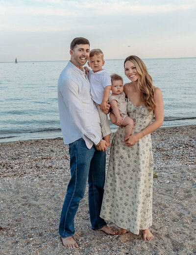 Professional family portrait by Stamford CT photographer featuring parents with two young children on a pebble beach at sunset, wearing coordinated neutral and blue tones