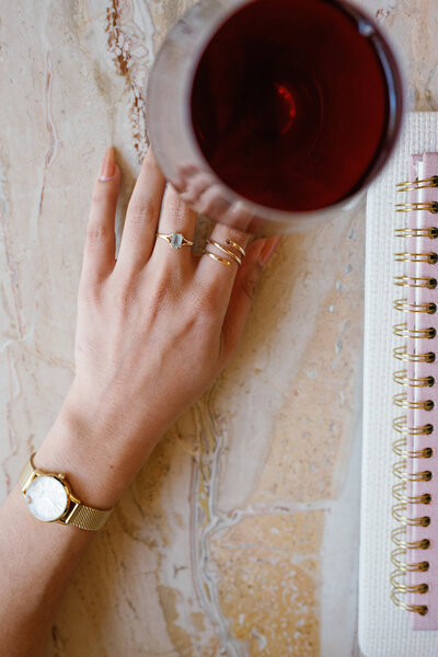 Woman's hand with a gold watch and rings resting next to a glass of red wine on a marble table