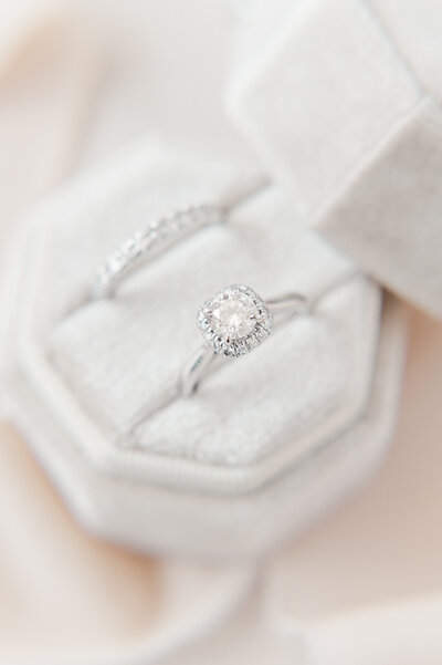 groom ring laying down on a flat white surface with brides ring propped up showing the stones in the engagement ring surrounded by flowers