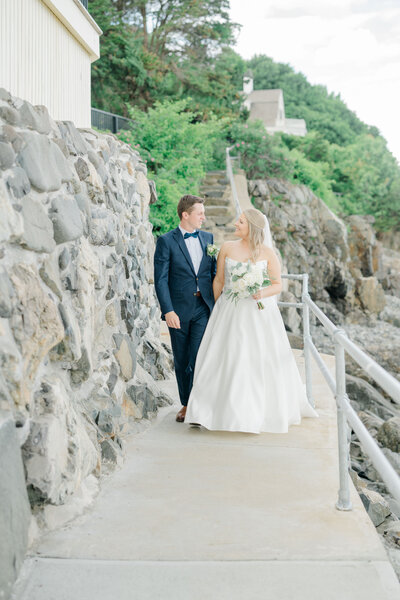 Wedding couple walking hand-in-hand along a coastal path near the Reading Room in York, Maine. The bride wears a flowing white gown, and the groom is dressed in a classic navy suit, both smiling and enjoying their moment together. Felisha, a destination wedding photographer based in New England, specializes in capturing timeless moments and scenic weddings in Maine, New England, and worldwide locations.