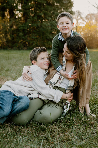 Mom sitting on the ground with one son hugging her on the left side and the other hugging her from behind around her neck in Gambrills  Maryland