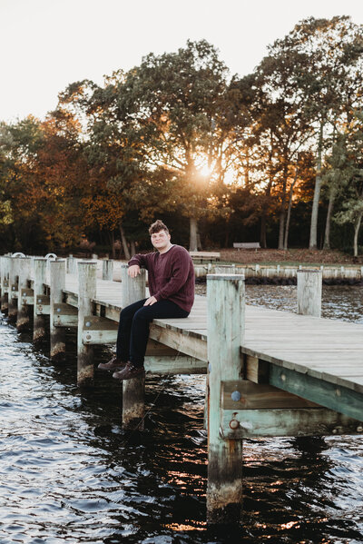 Teen boy sitting on pier in Annapolis Maryland wearing red sweater and black pants.