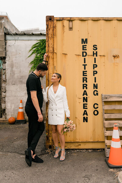 couple standing near a shipping container looking at each other as the woman rests her hand on the man's chest