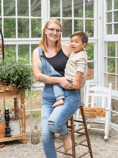 A mom wearing overalls holds her toddler. She is sitting on a stool inside a greenhouse.