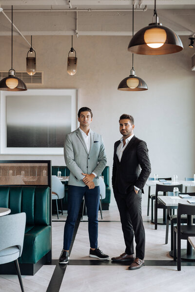 Two professionally dressed men pose confidently in a modern, upscale restaurant with contemporary decor. One wears a light gray blazer with navy pants, while the other wears a black suit with an open-collar white shirt. The setting features stylish green booth seating, elegant pendant lighting, and minimalist artwork on the walls.