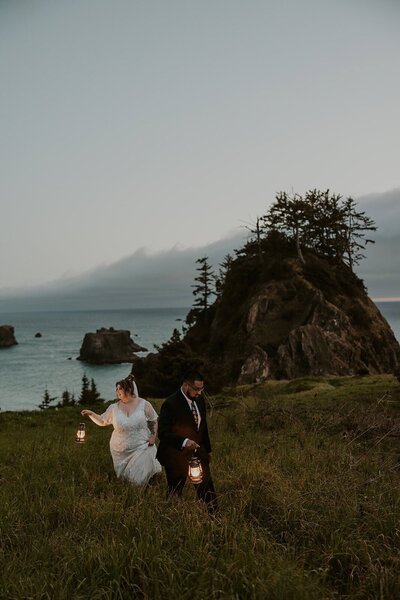 A couple walks on a grassy hill in wedding attire with vintage lanterns on the Oregon Coast